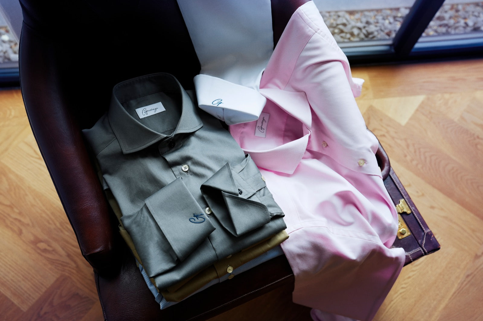 A curated stack of high-quality men's dress shirts in sage green, light pink, and white, draped over a leather armchair. The sage green shirt in the foreground features a prominent custom 