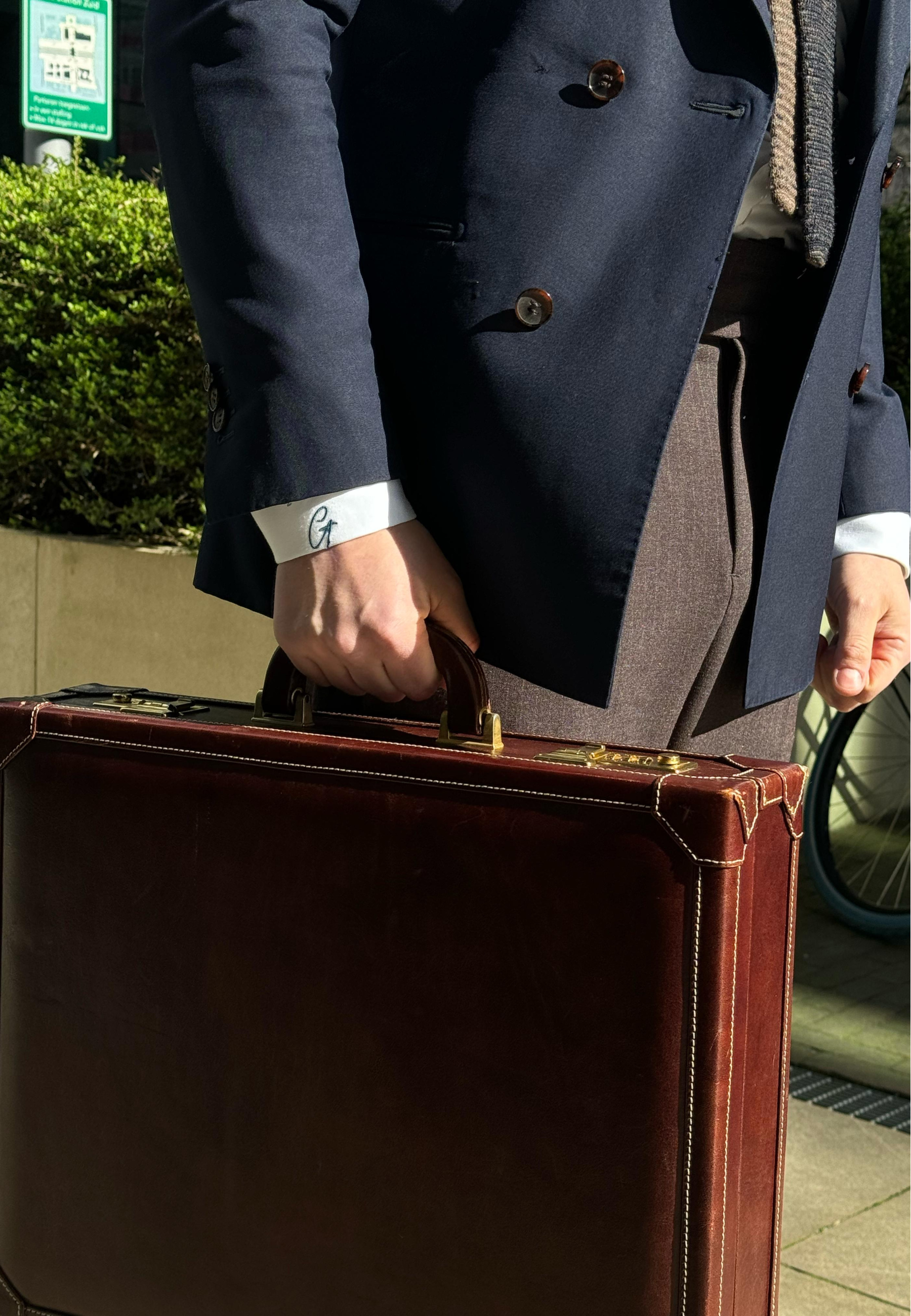 Person holding a brown suitcase with a suit jacket and watch visible

